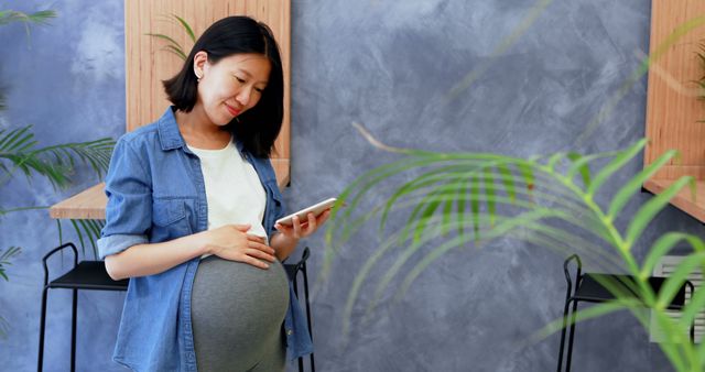 Pregnant woman standing in modern café, using smartphone, and wearing casual outfit with denim jacket and white shirt. Background includes sleek wooden furniture and lush green plants. Ideal for content related to motherhood, modern lifestyle, technology, and café culture.