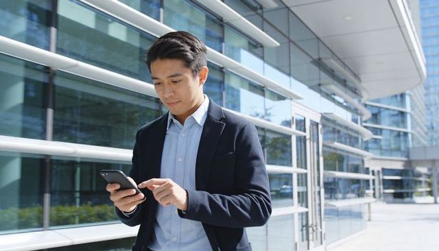 Asian businessman checking smartphone outside modern glass office building, professional