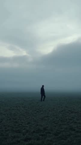 Vertical video of lone man walking across frost-covered plain through moody morning fog