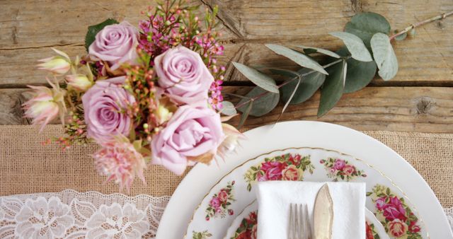 Top-down view of a rustic table decorated with a bouquet of pink roses, floral plates, and elegant lace. Ideal for use in wedding decor inspiration, vintage-themed parties, dining promotional materials, or farmhouse style decoration concepts.