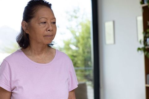 Senior asian woman standing by balcony door wearing pink t-shirt, looking reflective