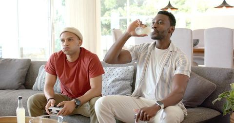 Friends gaming on couch while drinking bottled beverages, using controller and remote