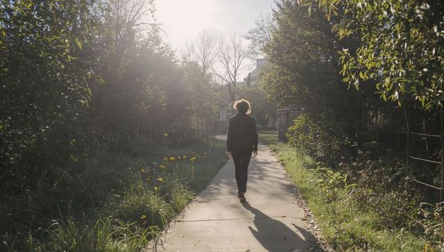 Backlit woman walking along sunlit suburban path through green foliage and wildflowers