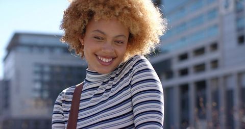 Sunlit smiling woman tilting head in striped turtleneck, urban backlit lifestyle portrait