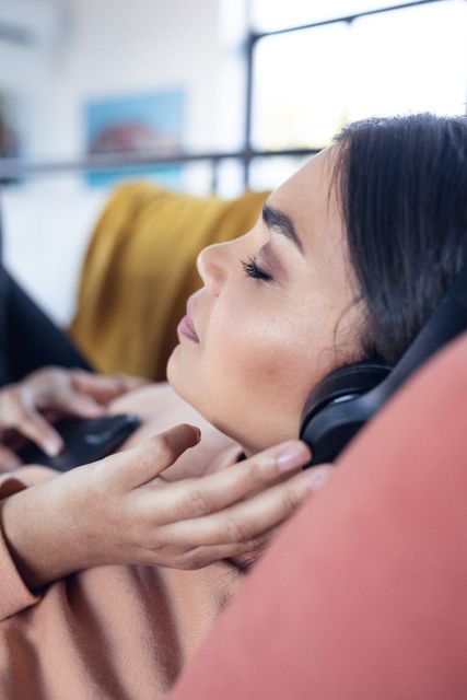 Vertical image of relaxed biracial woman lying on sofa and listening to music. Lifestyle, relax and spending free time at home concept.