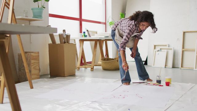 This scene captures an artist engrossed in her work, painting on a large sheet on the floor of a bright and airy studio. An appealing portrayal of the creative process, the workspace is scattered with various art supplies and materials, infusing spontaneity and innovation. This can be used in articles about the artistic process, creativity, or for promoting art workshops and classes focusing on artistic expression in unusual spaces.