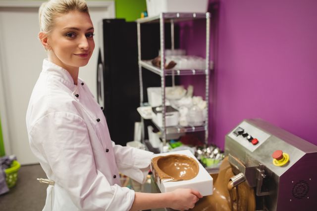 Female chocolatier pouring melted chocolate into a mould in a professional kitchen. Ideal for use in articles or advertisements related to culinary arts, chocolate making, confectionery, and professional chefs. Can also be used for content focusing on the food industry, dessert preparation, and sweet treats.