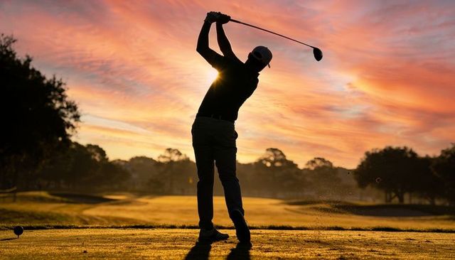 Silhouetted golfer swinging driver at sunrise over fairway with dramatic golden sky