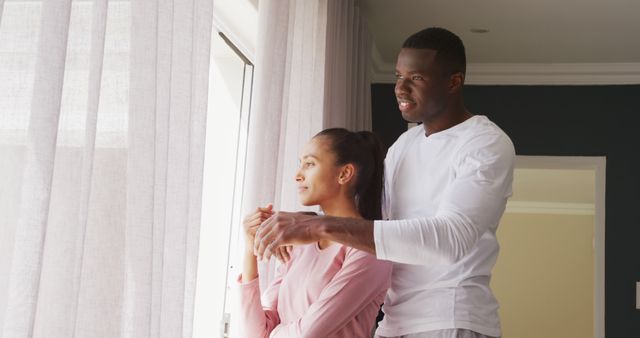 Image of happy diverse couple looking out of window and embracing at home. Happiness, love, domestic life, and inclusivity concept.
