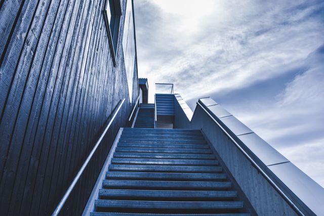 Upward view of metal and wooden staircase leading towards blue sky. Image captures clean lines and modern architecture, ideal for use in themes of progress, aspirations, or architectural features. Suitable for marketing materials, websites, and editorial publications highlighting contemporary design and construction.