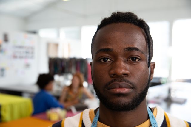 Portrait of a young African American male fashion student with short hair and dreadlocks in a studio at fashion college, wearing a striped sweater and a tape measure around his neck, standing and looking straight to the camera, with other students working in the background out of focus.