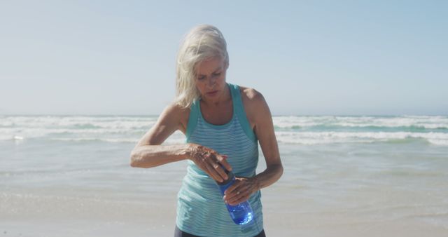 Senior woman seen drinking water while taking a break from her morning exercise on the beach with ocean waves in the background. Perfect for promoting healthy living, active lifestyles, hydration during workouts, or senior fitness programs.