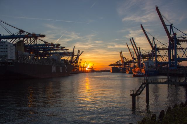 Industrial port at sunset featuring cargo ships and cranes lined up along the harbor, with their reflections mirrored in the water. This serene evening shot illustrates the intersection of natural beauty and industrial achievement. Suitable for use in backgrounds, transportation and logistics themes, and industrial and business contexts.