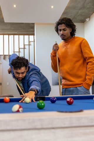 Indian friends playing pool in modern home rec room lining up precise shot on blue felt table