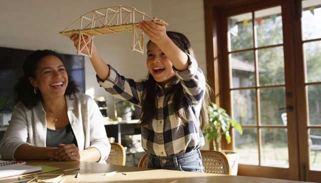 Child presenting handcrafted popsicle-stick bridge while parent smiling in sunlit home