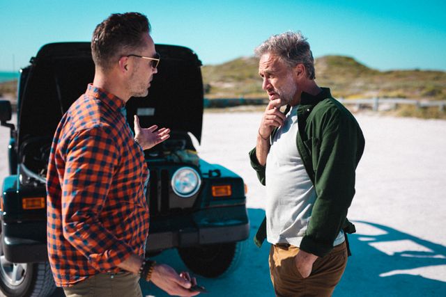 Two Caucasian men are seen discussing a car breakdown during a summer road trip. One man is wearing a plaid shirt and sunglasses, while the other is in a green jacket. The vehicle's hood is open, indicating a mechanical issue. This image is ideal for use in travel blogs, articles about road trip tips, automotive repair guides, and advertisements for roadside assistance services.