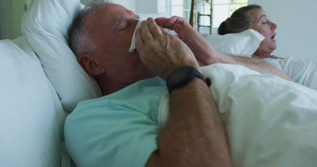 This stock photo captures a senior man lying in bed while blowing his nose, indicating he is not feeling well. His concerned partner looks on from the background. This image is ideal for illustrating articles or advertisements related to healthcare, illness recovery, elderly support, flu or cold symptoms, healthcare for seniors, and promoting products catering to elderly care.