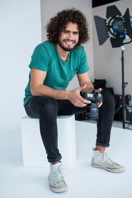Young male photographer with curly hair sitting on a white cube in a studio, holding a camera and smiling. He is dressed casually in a green shirt, black jeans, and sneakers. Ideal for use in articles about photography, creative professions, studio work, or lifestyle blogs.