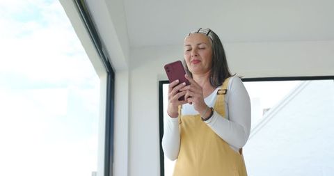Mature woman using smartphone in sunlit loft wearing mustard overalls and headband