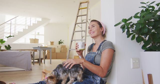 Woman taking a break while moving into a new home, sitting on the floor with a small dog, drinking from a cup. Ideal for representing concepts of moving, new beginnings, companionship, and home life. Useful in blogs, advertisements related to real estate, moving services, and lifestyle content.