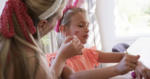 Mother painting daughter face with pink paint during playful home crafting and dress-up
