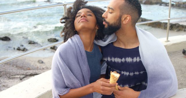 Young African American couple sharing a tender moment while enjoying ice cream by the ocean. They are wrapped in blankets, savoring their time together. Ideal for use in romance, vacation, lifestyle, and diversity themes.