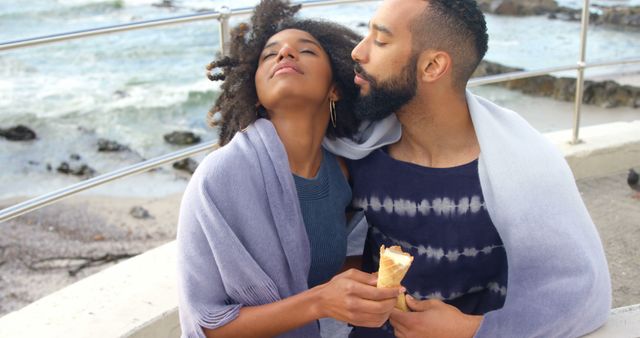 Romantic diverse couple with blankets on backs and ice creams embracing on sunny beach - Download Free Stock Photos Pikwizard.com