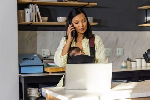 Indian mother juggling remote work and baby at kitchen island, talking on phone, using laptop