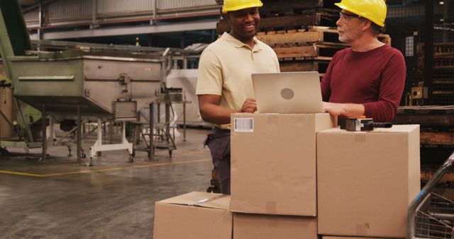 Two industrial workers with hard hats discussing logistics over packages in a warehouse environment. Ideal for use in articles about manufacturing, industry operations, warehouse logistics, teamwork, and productivity in industrial settings.