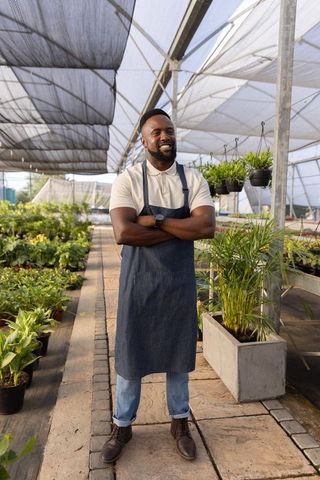 Smiling african american nursery owner standing in greenhouse wearing denim apron