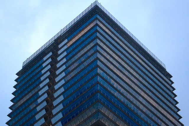 Angled view of a modern skyscraper with a geometric design against a blue sky. Useful for projects focusing on urban development, modern architectural styles, real estate, and commercial property promotion. Ideal for showcasing city life, business environments, and corporate culture in a contemporary setting.