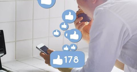 Asian man scrolling smartphone while eating cupcake in kitchen with social likes overlay