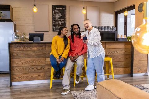 Diverse friends sharing smartphone and laughing at cozy coffee bar with yellow stools