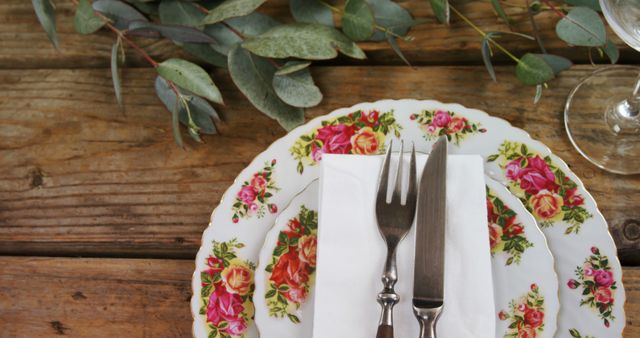 Elegant table setup featuring floral patterned plates with a knife and fork on a white napkin, placed on a rustic wooden table decorated with eucalyptus leaves. Suitable for content related to dining, table decor, restaurant settings, or home entertaining.