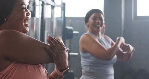 Smiling diverse mid-adult women stretching in gym wearing fitness tracker and activewear