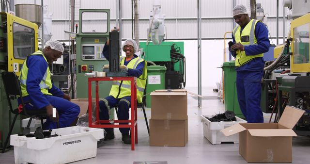 Three factory workers laugh and take a break in an industrial setting. They wear safety gear, including hi-visibility vests and hard hats. They are surrounded by machinery and open cardboard boxes, emphasizing an active production area. This image can be used to depict workplace camaraderie, safety practices, or team building in an industrial or manufacturing environment.