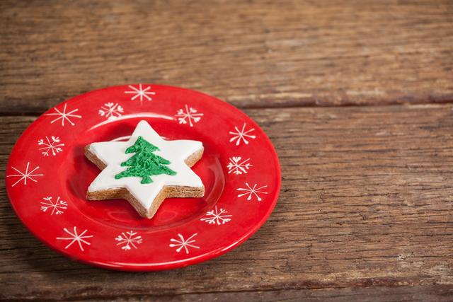 Star-shaped Christmas cookie with green tree icing on red festive plate with snowflake design. Wooden table background adds rustic charm. Ideal for holiday-themed promotions, festive greeting cards, and seasonal recipe blogs.