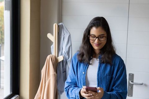 Young asian woman using smartphone and smiling in sunlit hallway wearing glasses and denim