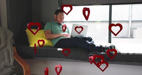 Man reclining using laptop on window seat with floating hearts ar overlay home workspace