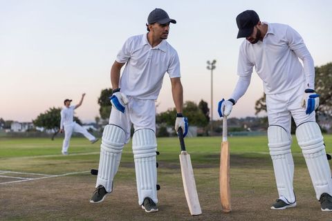 Diverse cricket teammates standing on pitch preparing to bat at dusk in pads and gloves