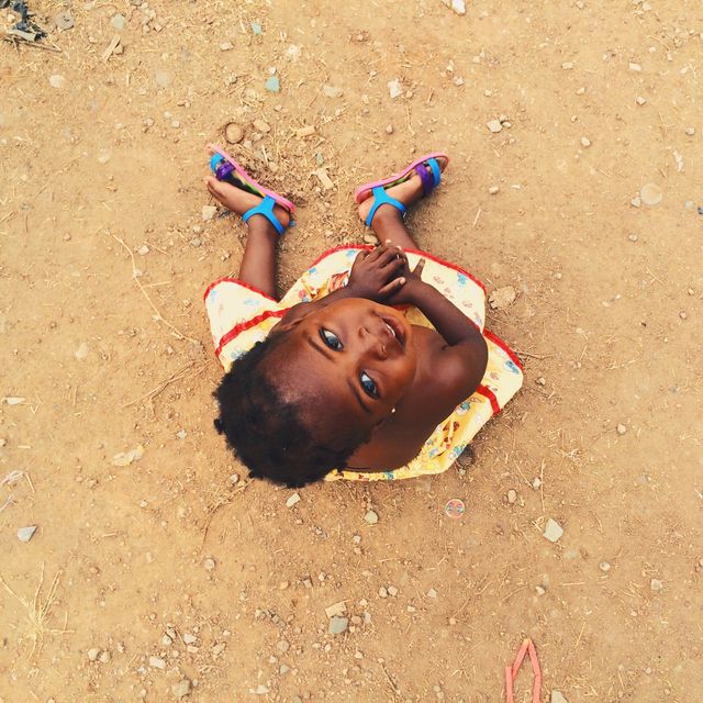 Smiling child wearing colorful clothing sitting on a dirt ground looking up. The image captures a candid moment of childhood happiness and innocence, making it ideal for themes related to joy, freedom, and rural life. Perfect for use in campaigns promoting children's welfare, education, and joyful outdoor activities.