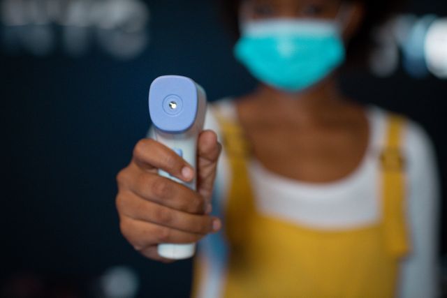 This image depicts an African American woman wearing a face mask and using an infrared thermometer to check temperature in an office setting. It is ideal for illustrating workplace safety measures, health precautions, and COVID-19 related protocols. Suitable for articles, blogs, and informational content about pandemic safety, office health guidelines, and protective measures in professional environments.