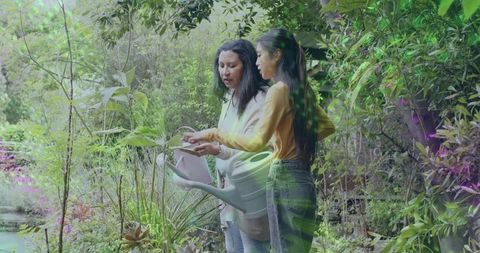Two women tending lush backyard garden reading plant tag holding white watering can