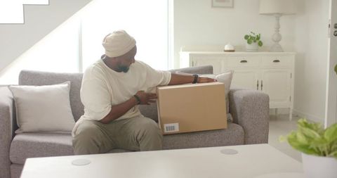 African american man receiving delivery box while sitting on sofa in bright minimalist living room