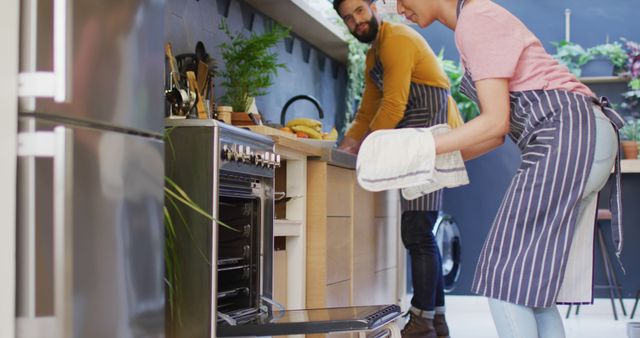 Image of happy diverse couple in aprons baking together in kitchen, using oven, with copy space. Happiness, communication, inclusivity, free time, togetherness and domestic life.