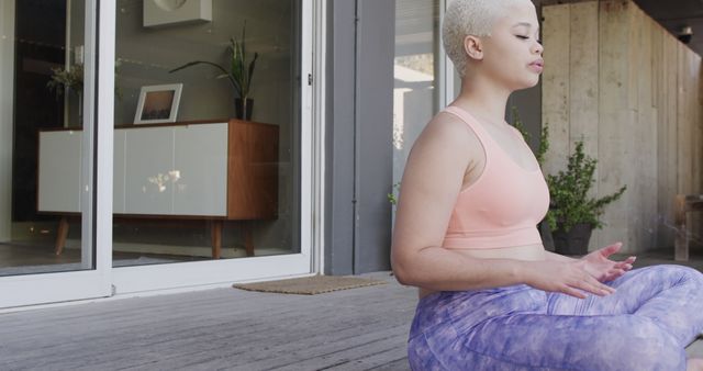 Young woman with short hair practicing mindfulness meditation on patio. She is seated in a lotus position, wearing a light pink sports bra and purple leggings. The scene has a modern, calm ambiance, suitable for wellness, self-care, outdoor activities, and fitness-related uses. This can be used in blogs, articles, social media posts, and promotional materials to highlight mental health, yoga practices, or personal wellness journeys.