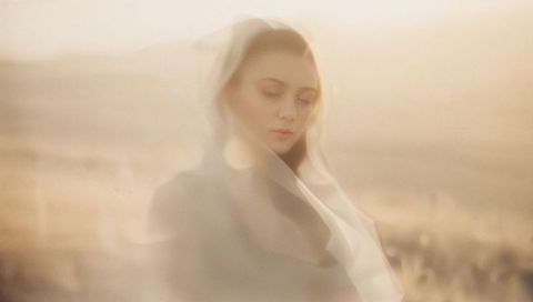 Standing woman in veil gazing downward in golden meadow at sunset soft-focus portrait