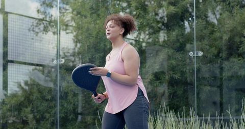 African american woman playing paddle tennis on glass court wearing pink activewear
