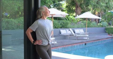 Senior woman standing by pool deck wearing smartwatch enjoying sunlit backyard relaxation