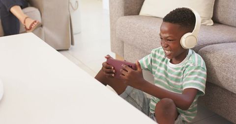 Young boy gaming on smartphone with headphones in cozy living room, grinning and focused